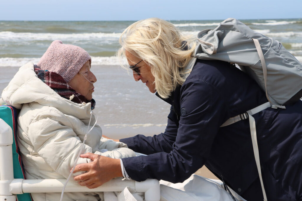 Frau beugt sich herunter zu einer kranken Frau im Rollstuhl am Meer